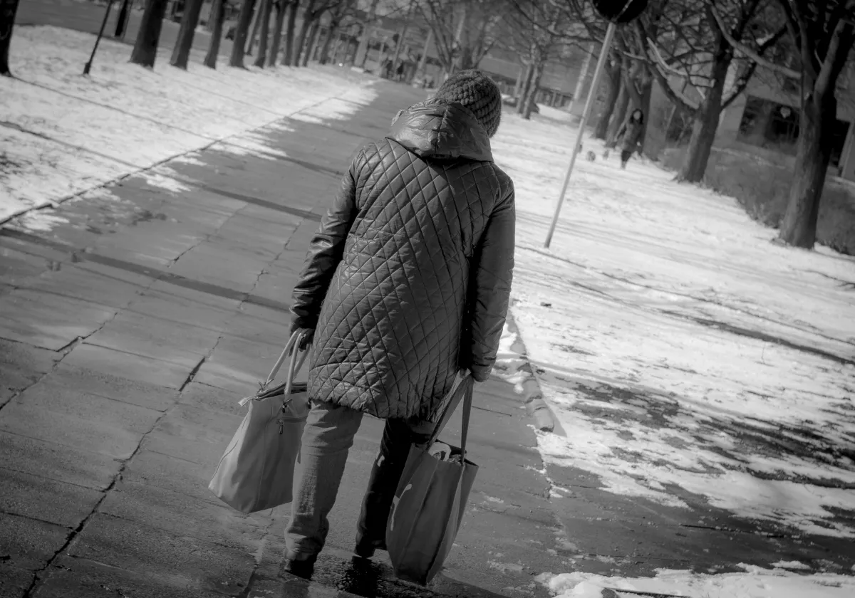 Woman in a quilted coat carrying two shopping bags along a snowy sidewalk